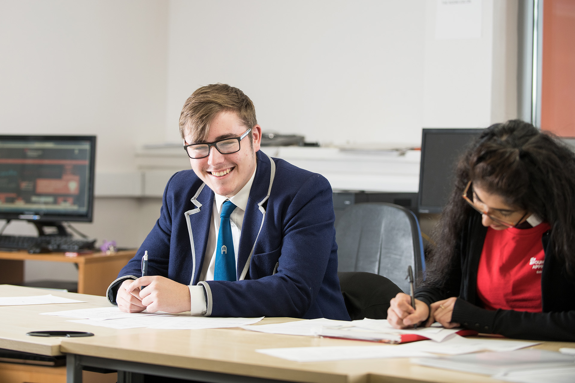 Young boy in a blue school uniform blazer and shirt and tie looks directly at the camera and smiles. He holds a pen in his hand. A young female is sitting next to him writing in her notepad.