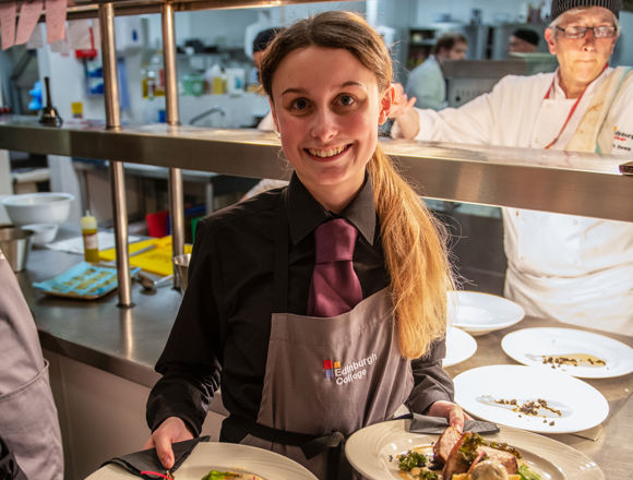 A waitress in a dark shirt and apron holds two elegantly plated dishes in a busy restaurant kitchen, with chefs working in the background.