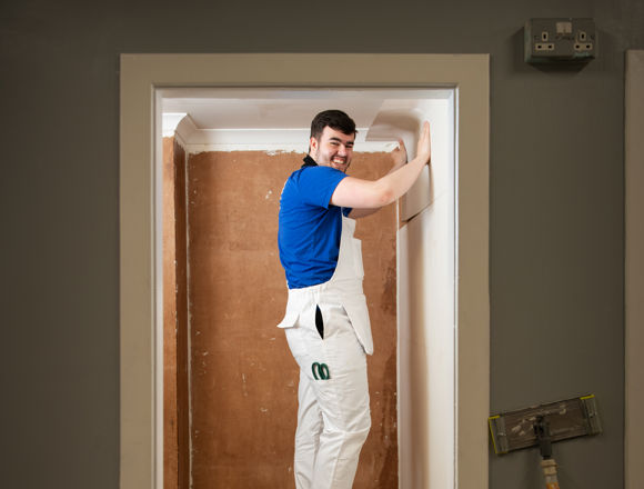 A person stands on a step ladder, applying finishing touches to a wall in a partially renovated room with a bare, brown wall.
