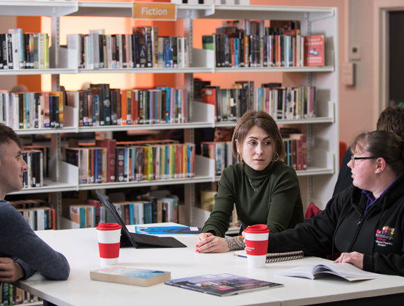 Three students working together at a desk in the library, with book shelves surrounding them.