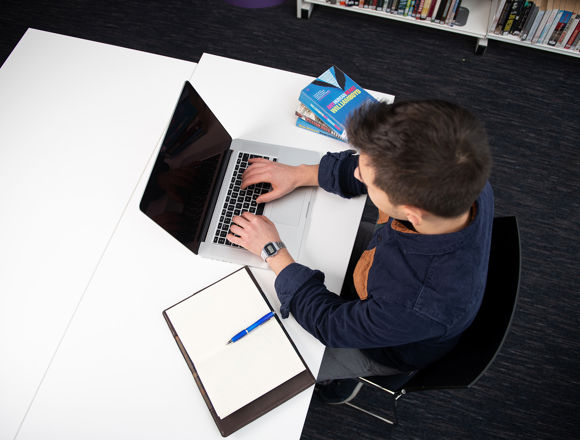 Student typing at their laptop in the college library, with books surrounding them.