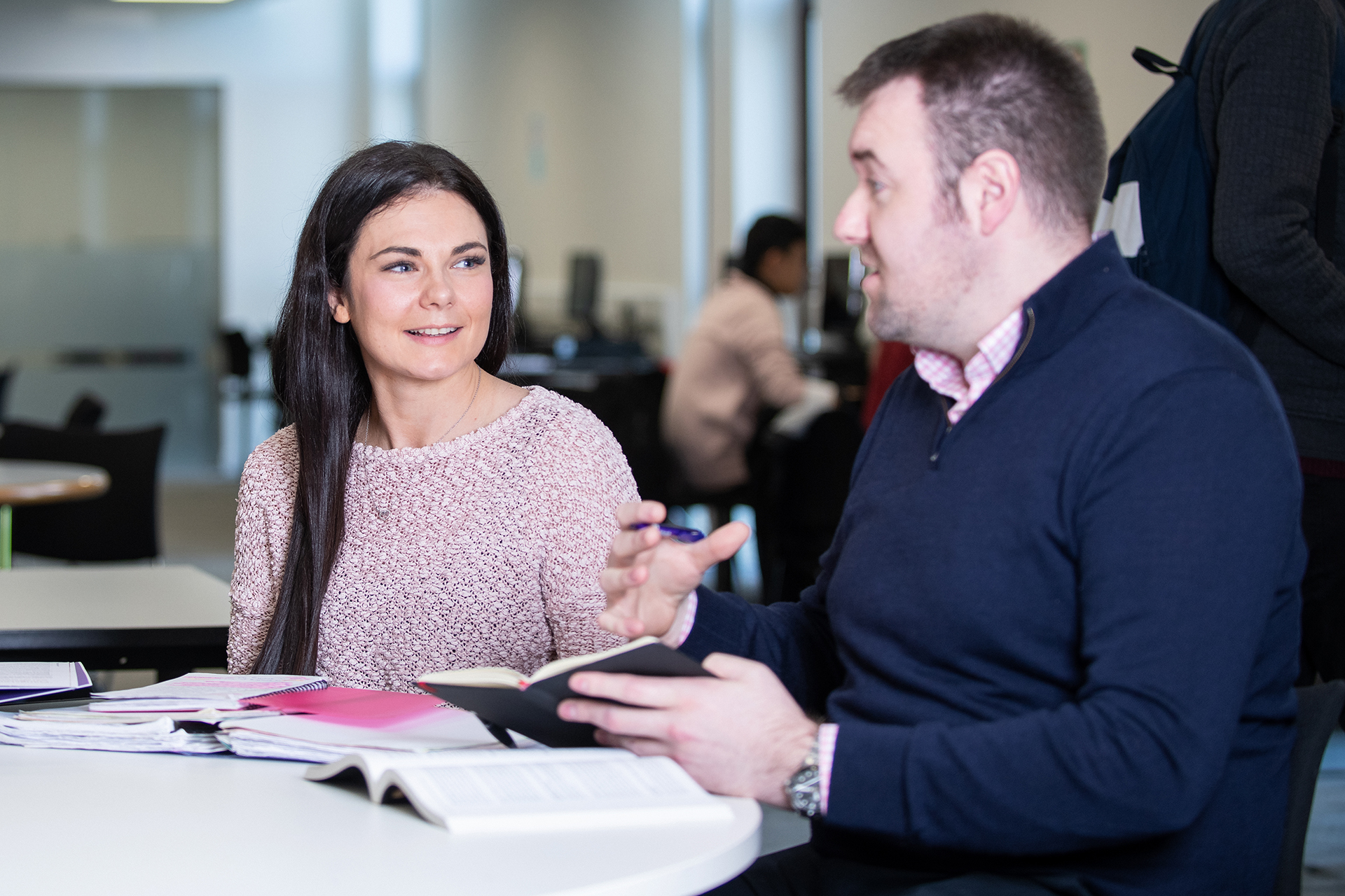 Student explaining something to another student while holding a notebook.