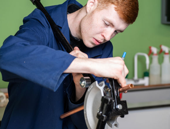 A person in a blue work jacket is bending a copper pipe with a pipe bender tool, focusing intently on the task.