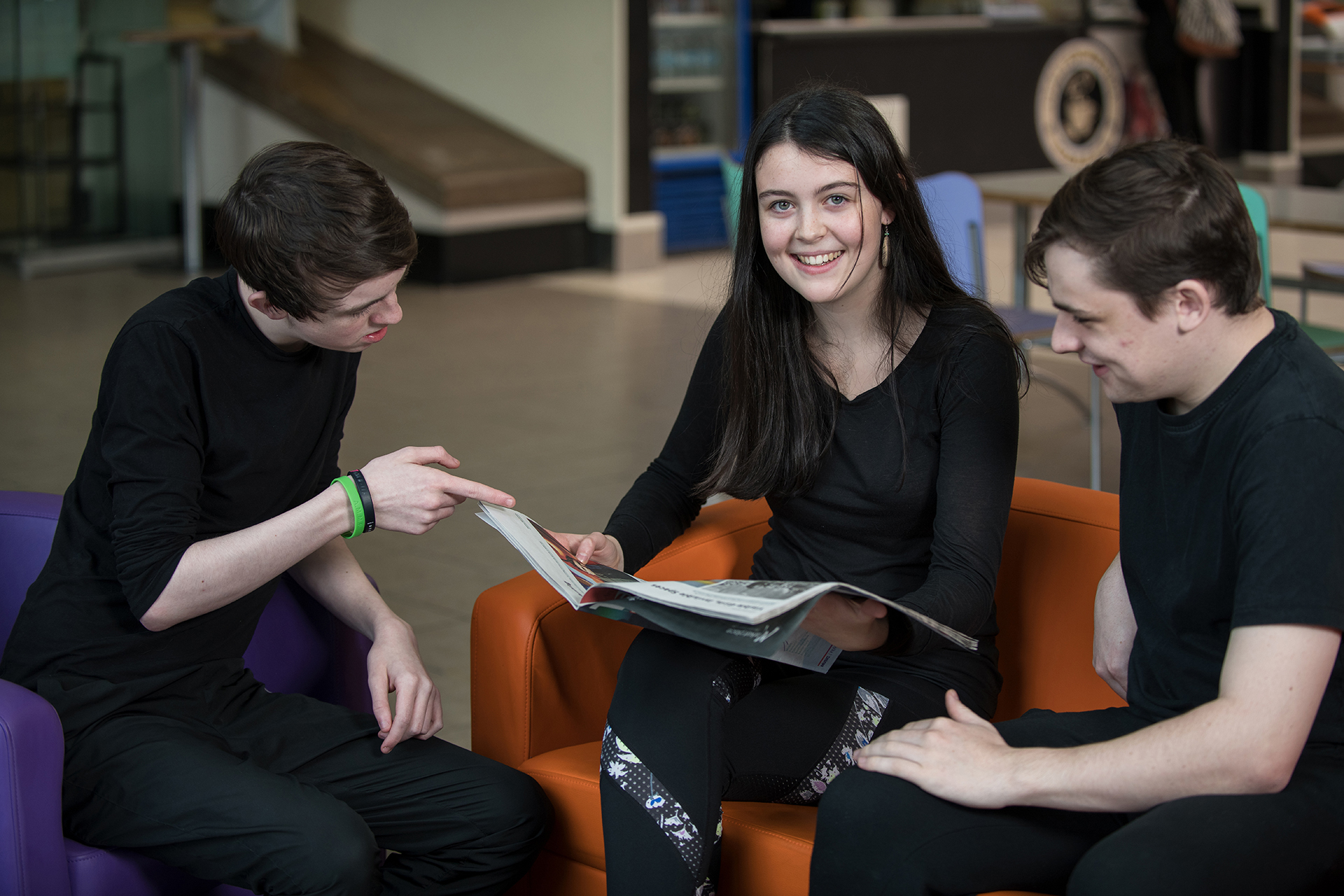 Two males looking at a book together with a female sitting in the middle looking directly at the camera and smiling.