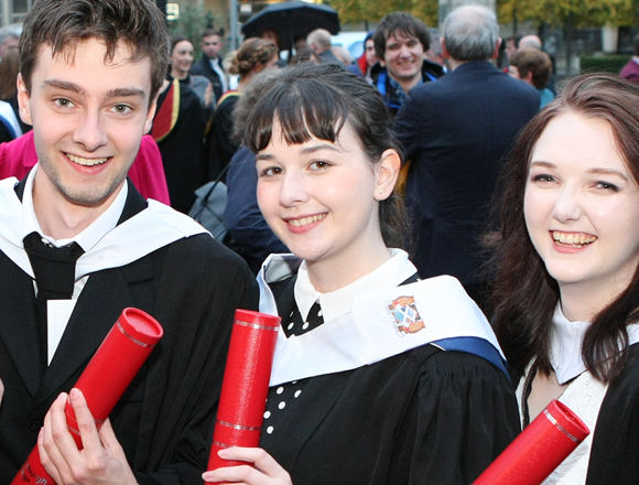 Four students at graduation, wearing black graduation gowns and holding red tubes with their scroll in.