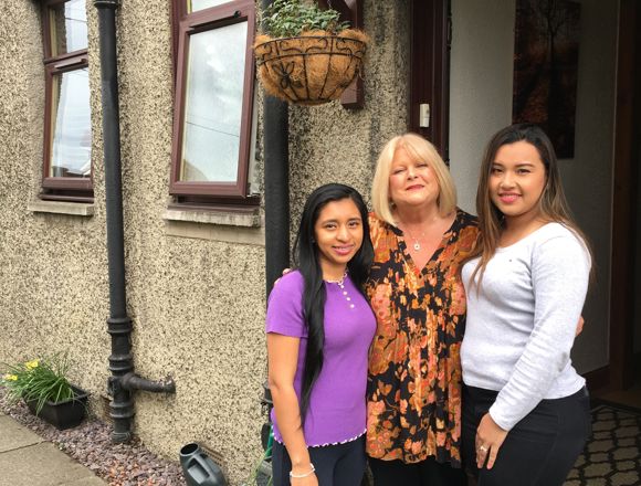 Three women stand together outside a textured house, with a hanging plant above and garden accessories visible on the ground.