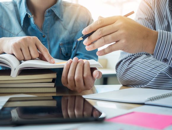 Two students are pointing at something in an open textbook, on top of a stack of closed textbooks.