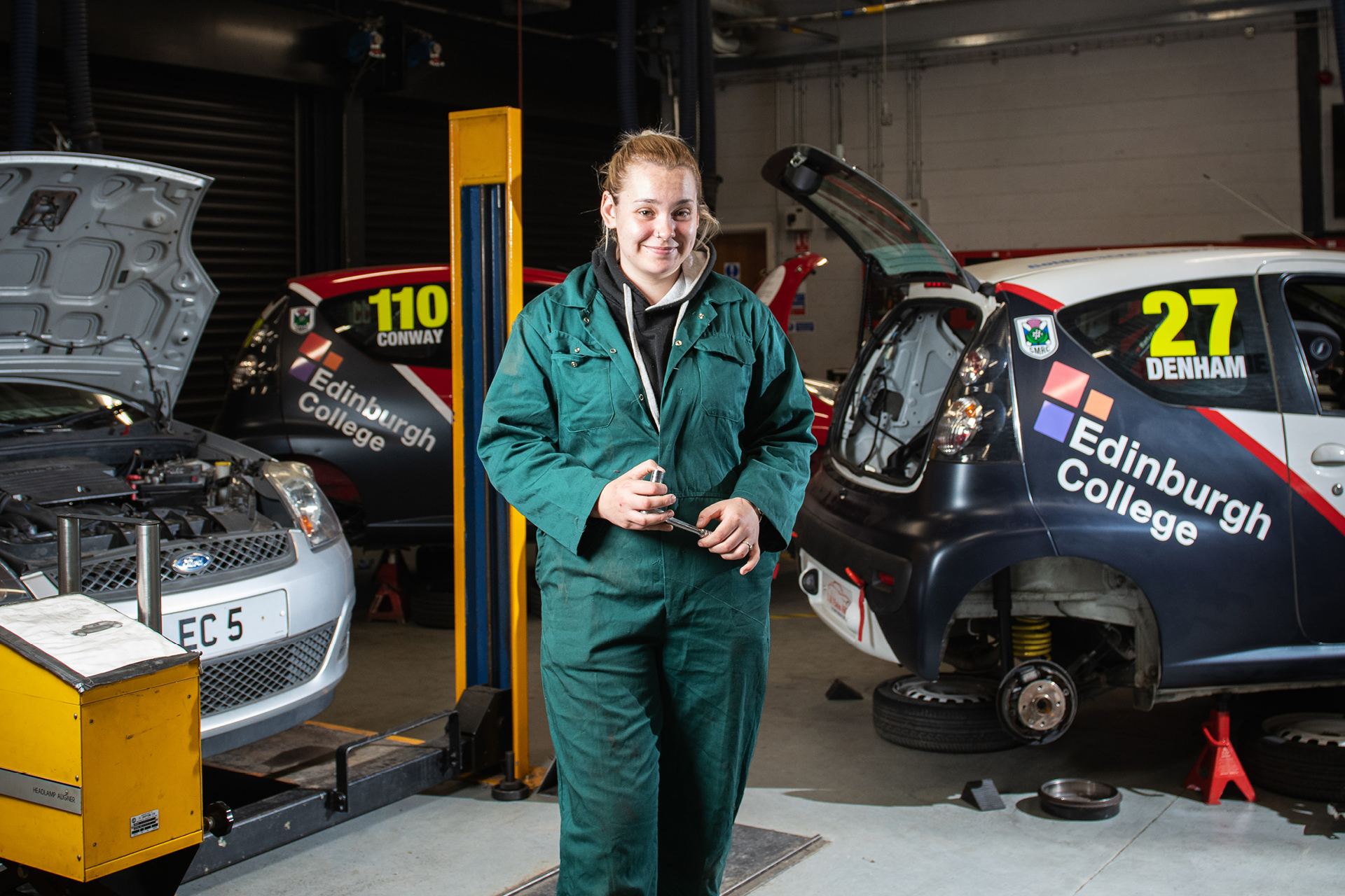 Automotive student standing in a garage with two cars getting worked on in the background.
